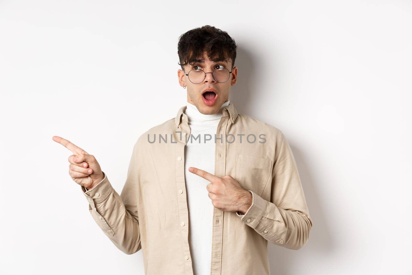 Amazed young man in glasses drop jaw, saying wow, pointing and looking left at awesome promo offer, stare with disbelief and impressed face, white background.
