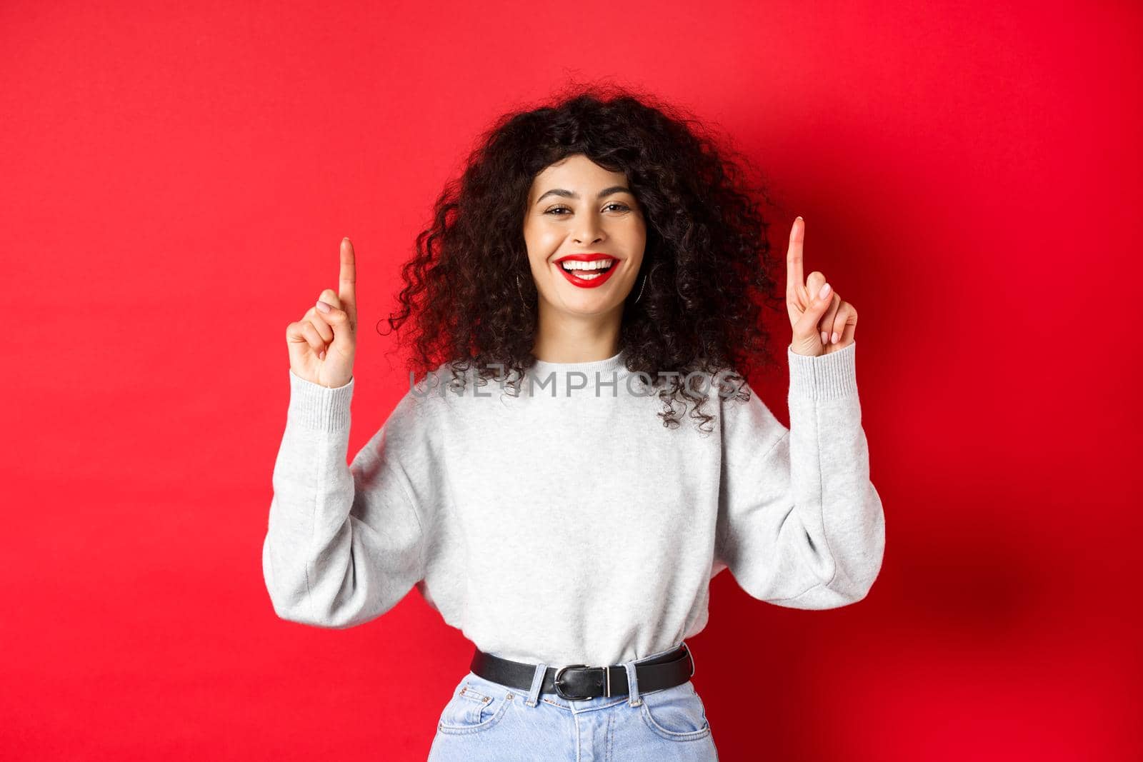 Happy young woman with curly hair, pointing fingers up and laughing, showing perfect white smile, standing against red background.