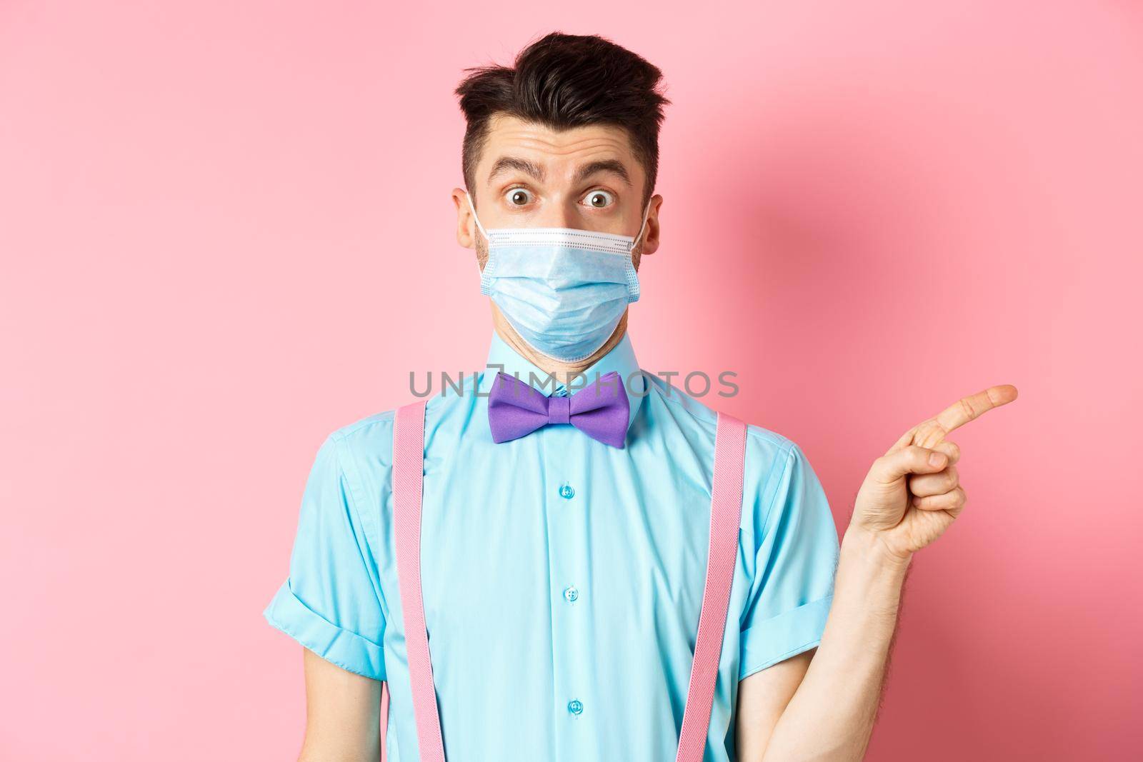 Coronavirus, healthcare and quarantine concept. Man looking surprised in medical mask, asking question and pointing right, curious about promo offer, standing on pink background.