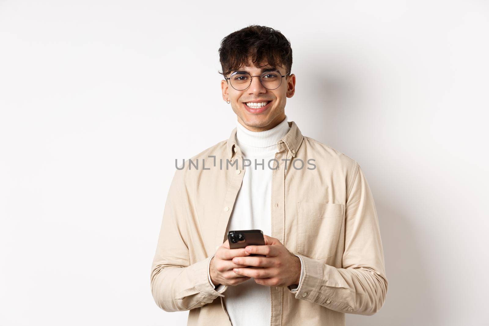 Real people. Handsome guy in glasses using mobile phone and smiling happy at camera, standing on white background.