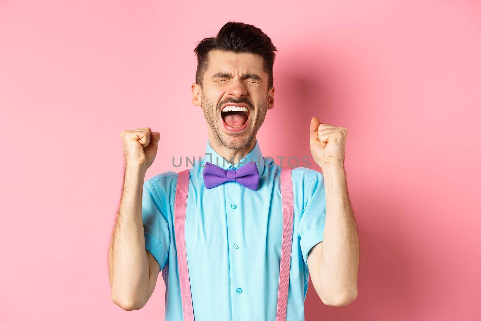 Relieved man shouting from happiness and joy, scream yes with closed eyes and clenched fists, celebrating victory, achieve goal and triumphing, standing over pink background.