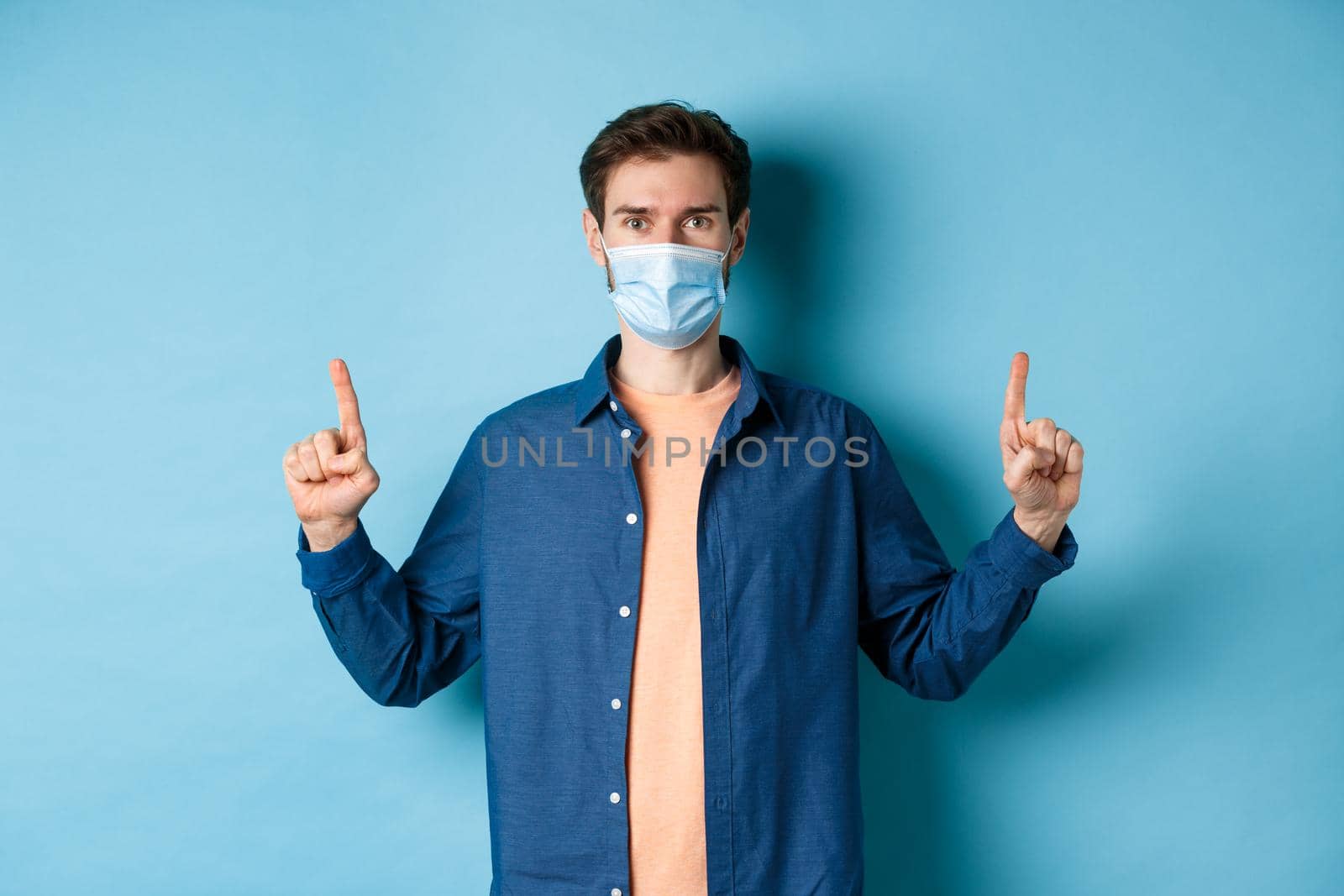 Covid-19 and lifestyle concept. Handsome young man in face mask looking healthy and happy, pointing fingers up at empty space, blue background by Benzoix