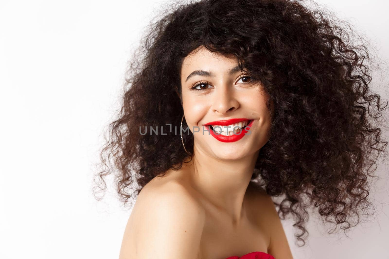 Close-up of cheerful smiling woman with red lipstick and curly hairstyle, looking happy, standing over white background by Benzoix
