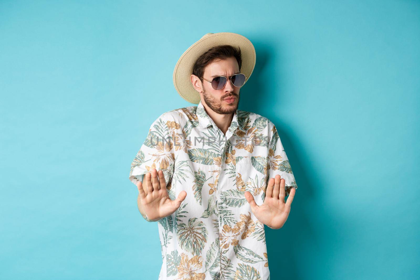Alarmed tourist asking to stay away, step back from something cringe, showing rejection gesture, standing in straw hat and hawaiian shirt, blue background by Benzoix