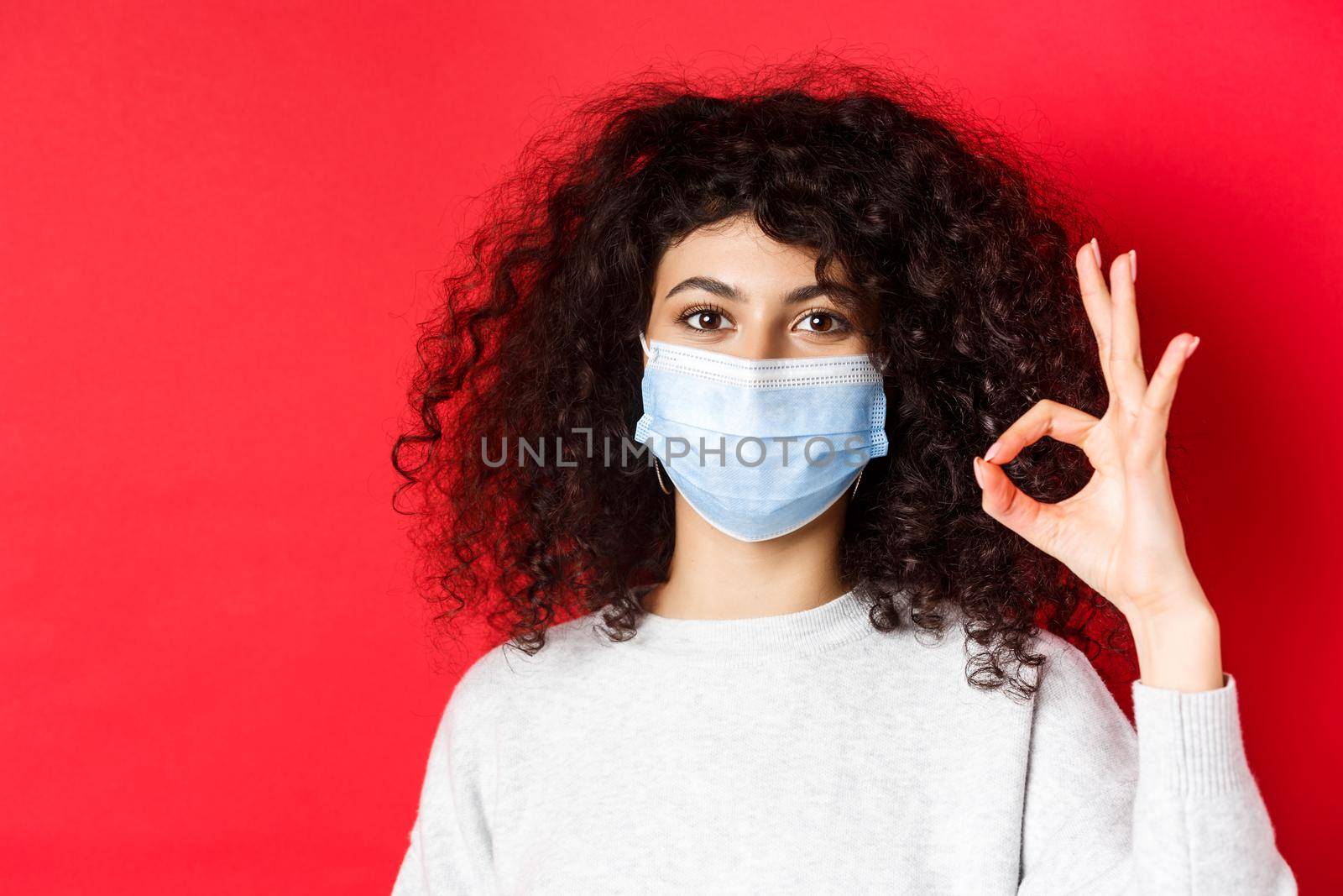 Covid and health concept. Confident young woman in medical mask showing OK gesture in approval, guarantee quality, standing on red background by Benzoix