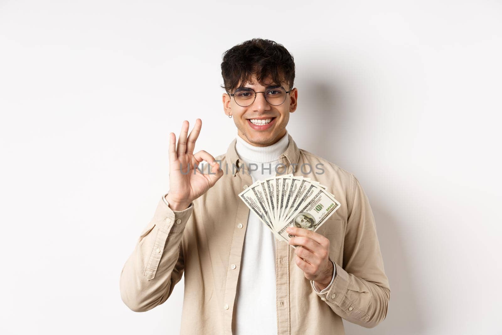 Handsome modern man in glasses showing dollar bills and okay gesture, making money, standing with cash on white background.