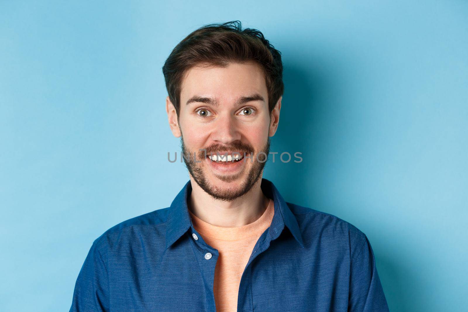 Close up of surprised handsome guy with beard looking amused, smiling happy at camera, standing on blue background.