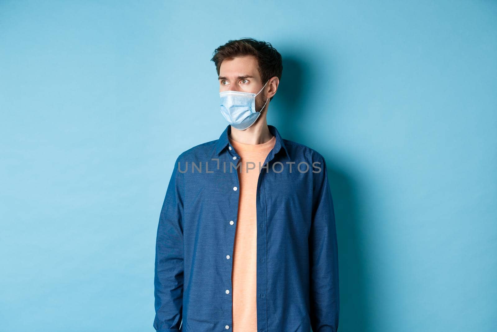 Covid-19 and healthcare concept. Shocked young man in medical mask looking aside at empty space on blue background by Benzoix