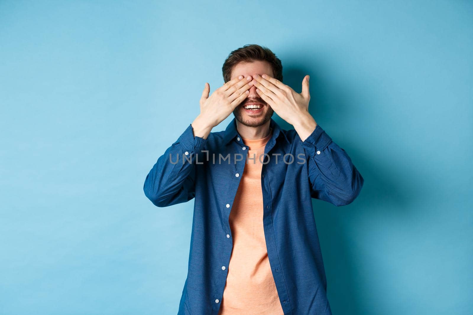 Happy smiling guy waiting for surprise, covering eyes with hands and anticipating gift, standing on blue background.