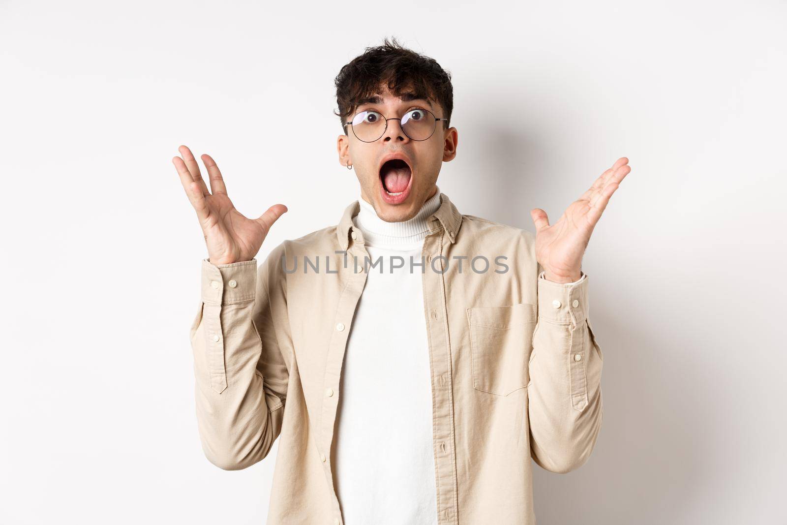 Scared young man in panic, screaming and looking axious, jumping startled and shocked, shaking hands nervously, standing on white background.