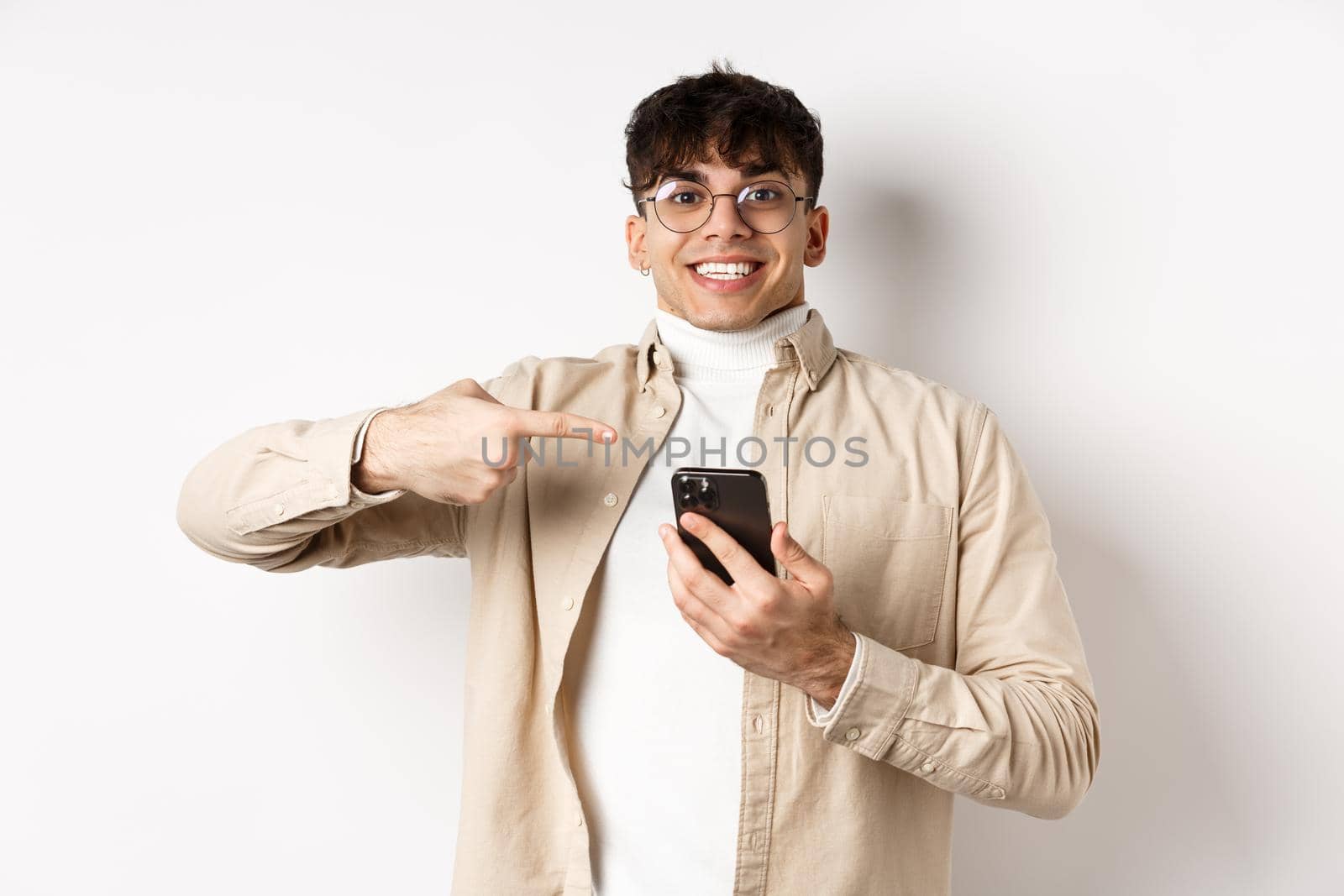 Technology and online shopping concept. Cheerful young man in glasses showing on mobile phone, pointing at smartphone screen and smiling excited, white background.