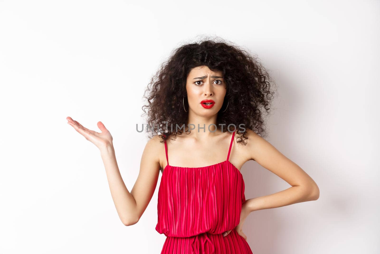 Upset pretty woman with curly hair and red dress, raising hand and look confused, frowning disappointed, talk about unfair things, standing against white background.