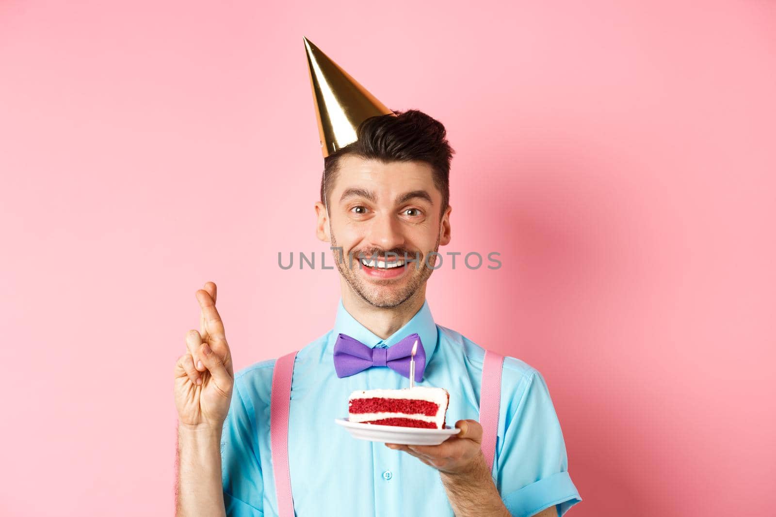 Holidays and celebration concept. Happy young man enjoying birthday party, wearing cone hat and cross fingers, making wish on bday cake with candle, pink background.
