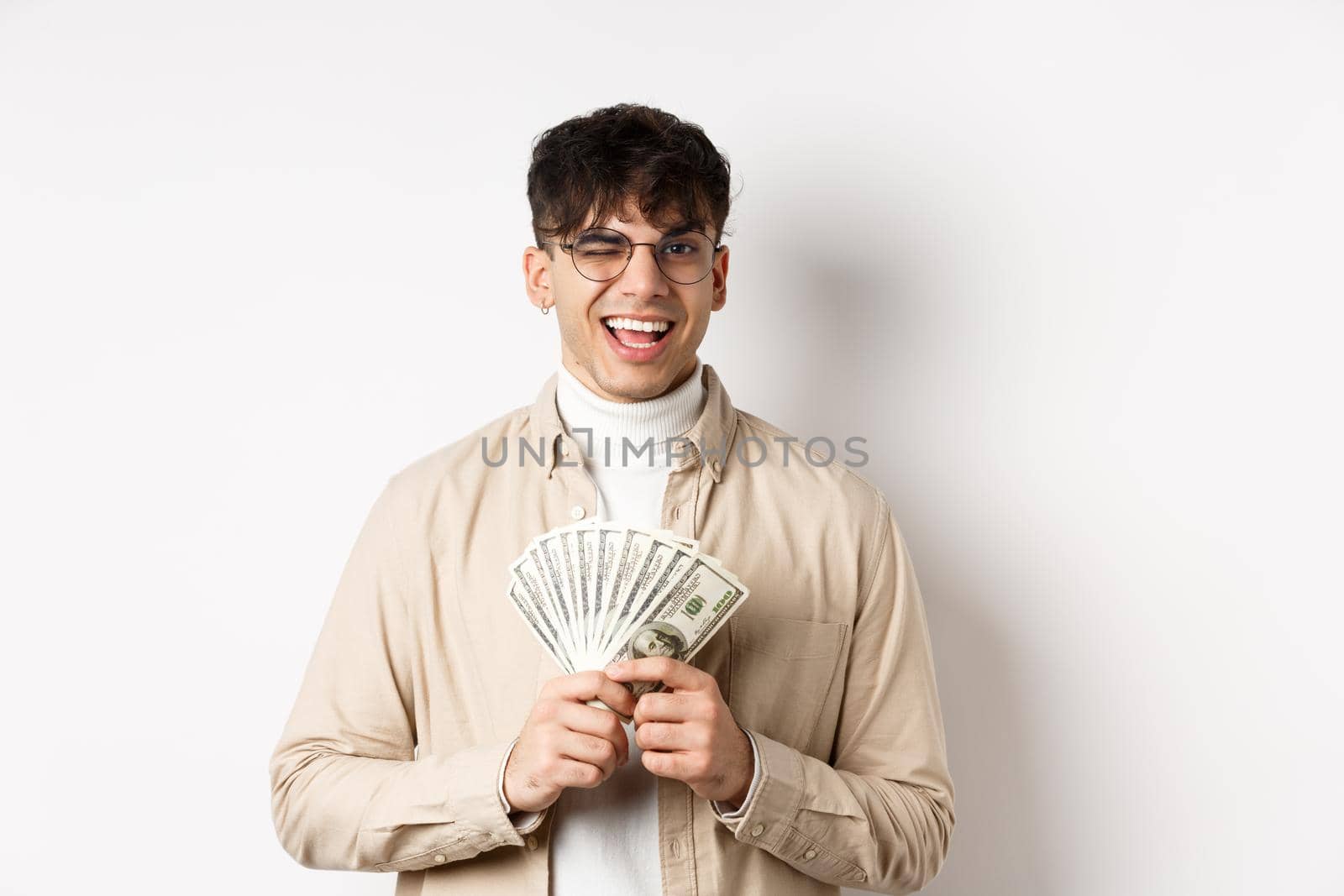 Handsome young man winking and showing dollar bills, holding fan of money banknotes and smiling pleased, making money, standing on white background by Benzoix