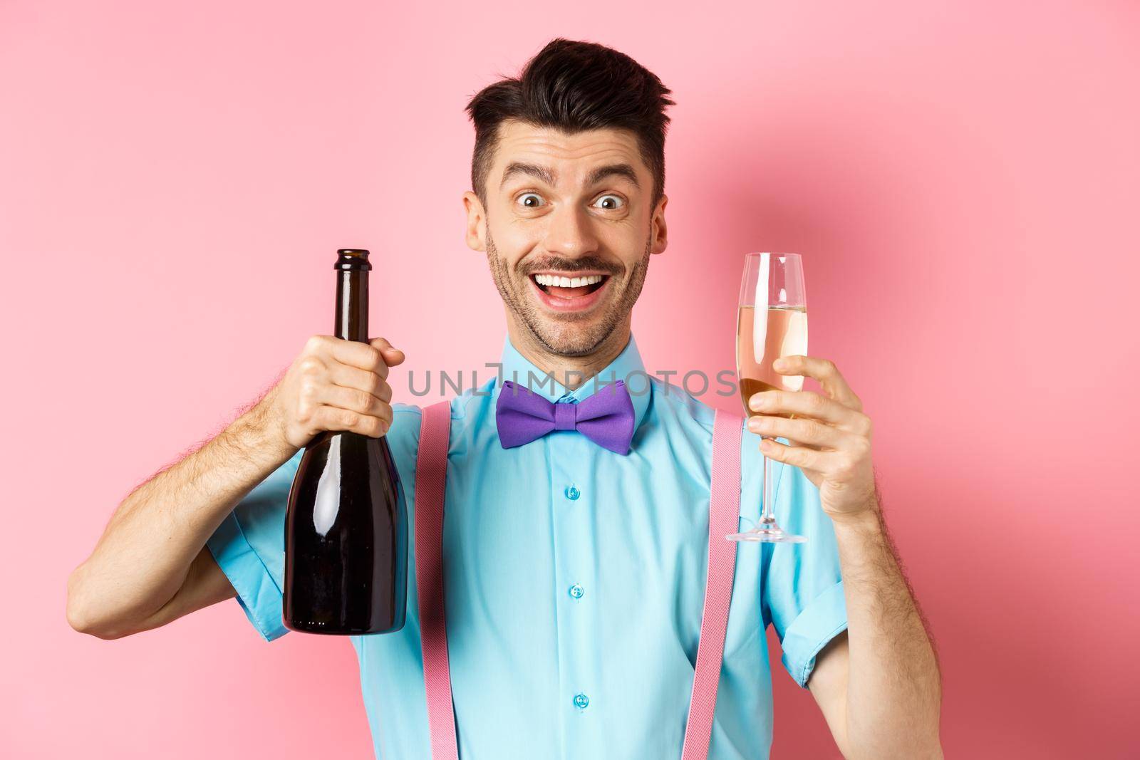 Holidays and celebration concept. Happy young man having fun, showing bottle of champagne and glass, making toast on event, smiling and looking at camera, pink background.