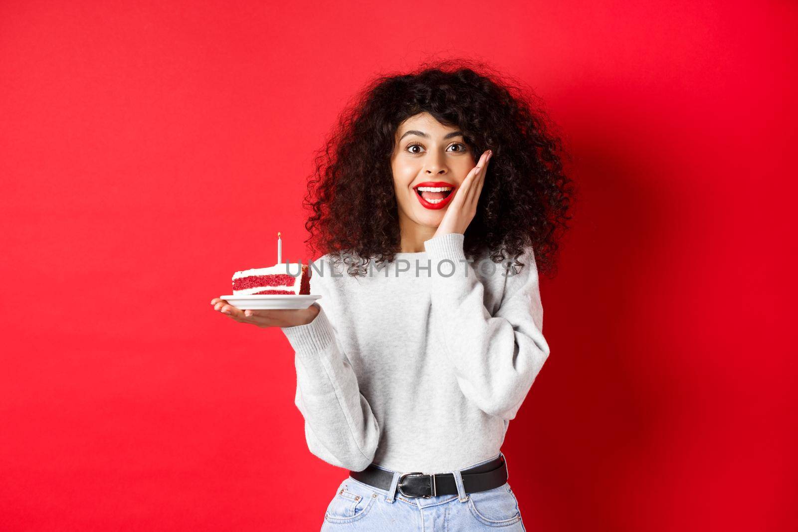 Excited woman celebrating birthday, holding cake and looking surprised and happy at camera, red background.