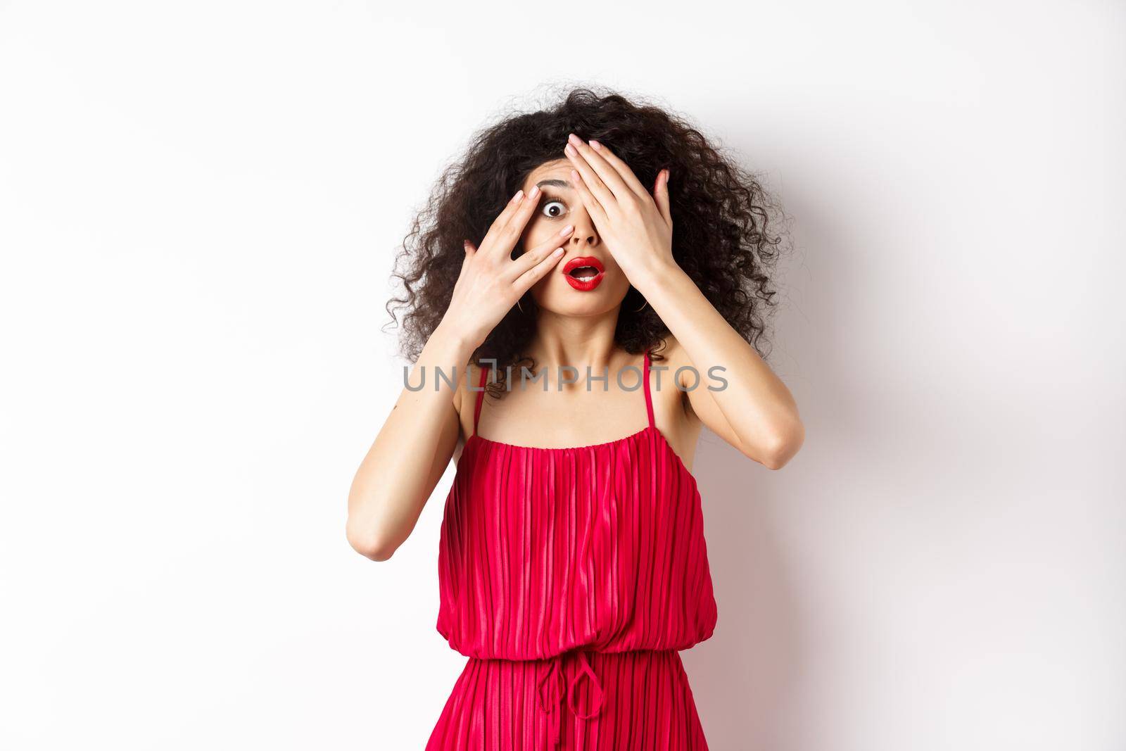 Excited curly-haired girl in dress, cover eyes with hands and peek through fingers, gasping shocked at camera, standing on white background.