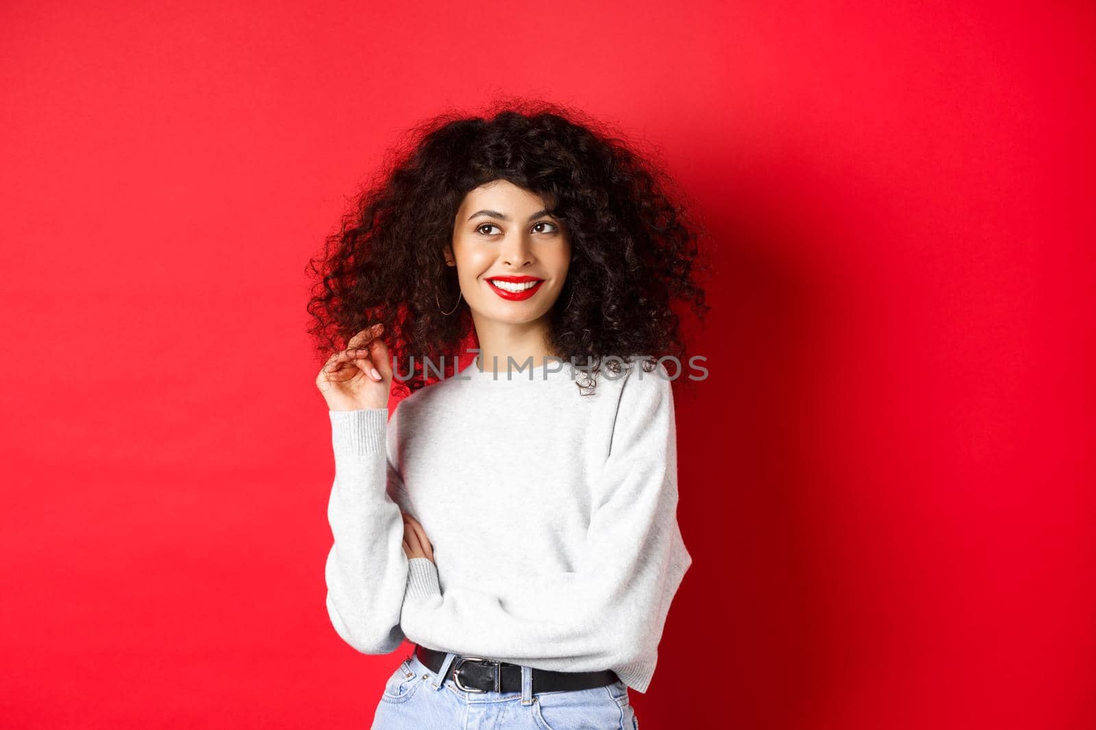 Modern woman in sweatshirt, playing with curly hair and looking aside at empty space, standing on red background by Benzoix