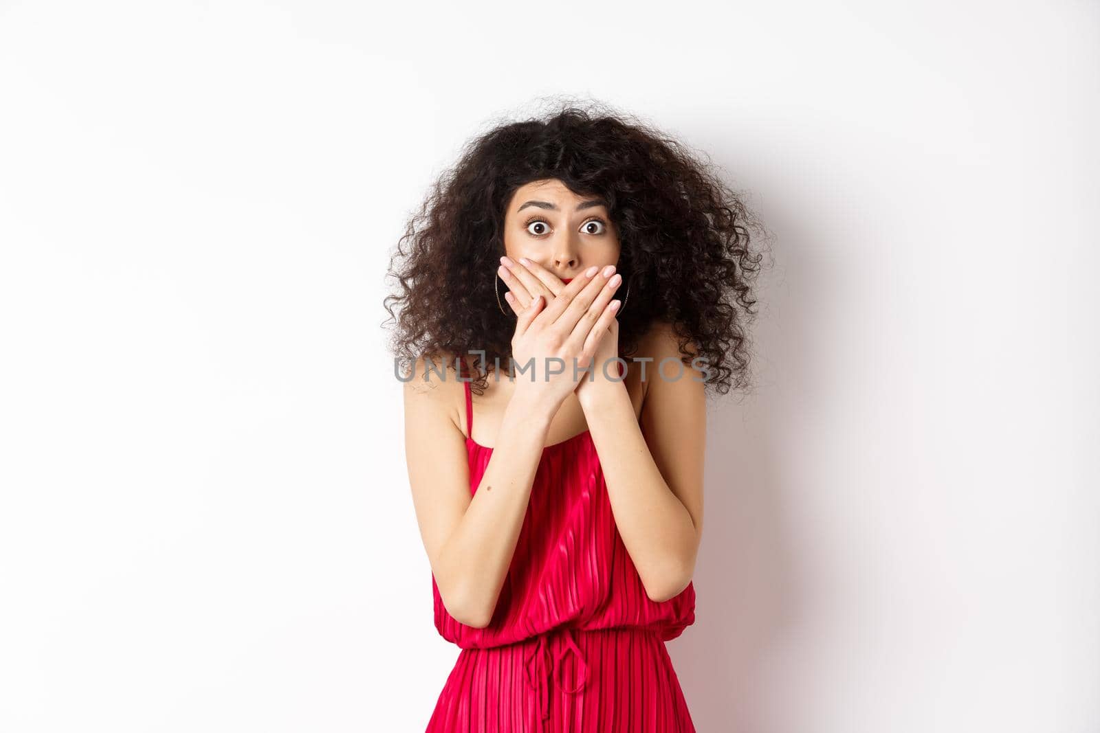 Shocked woman covering mouth with hands and staring with disbelief at camera, witness something shocking, standing in red dress on white background.