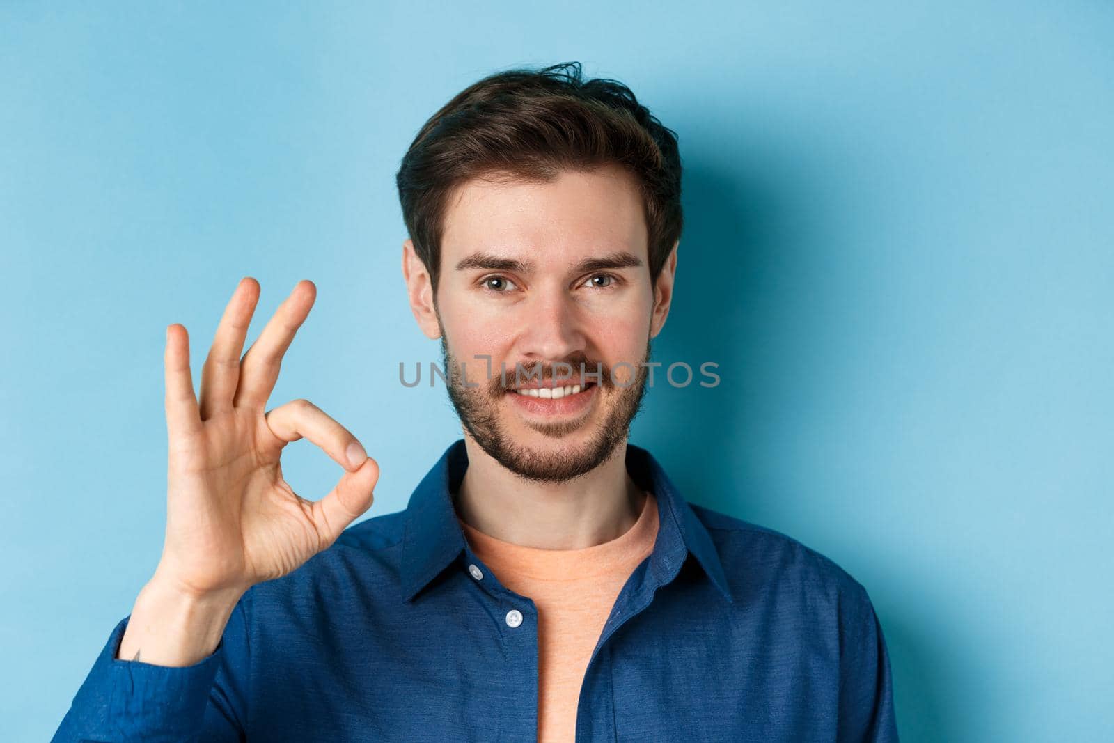 Close up of handsome bearded guy showing okay gesture and smiling, standing on blue background.