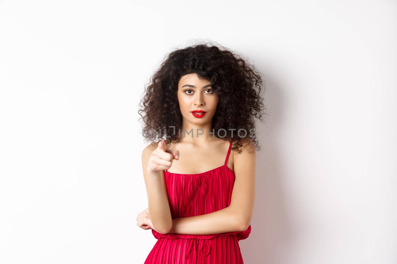 Confident young woman in red dress and makeup, pointing at camera, choosing you, inviting to event, standing over white background by Benzoix