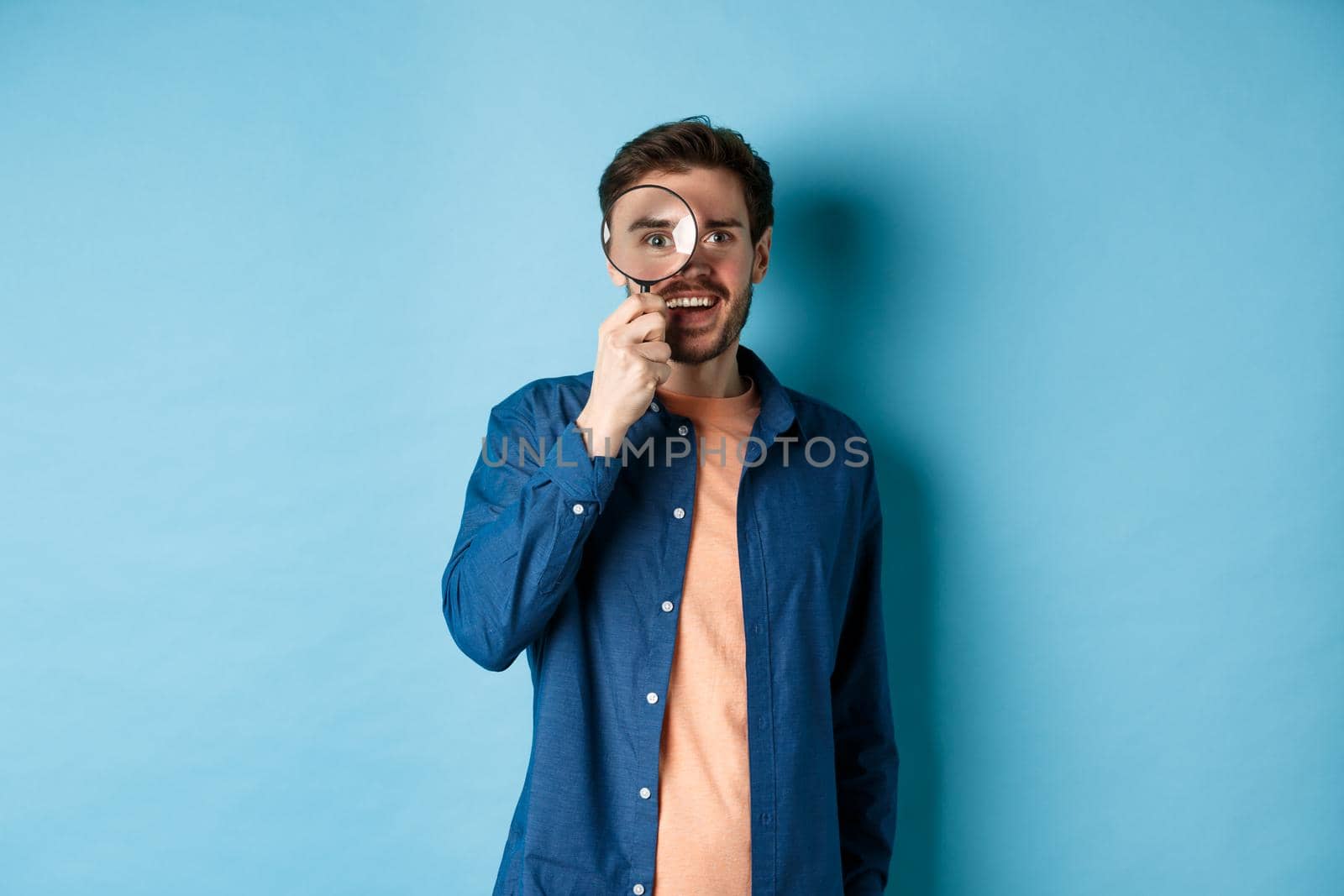 Cheerful young man found something good, smiling and looking through magnifying glass, standing on blue background.