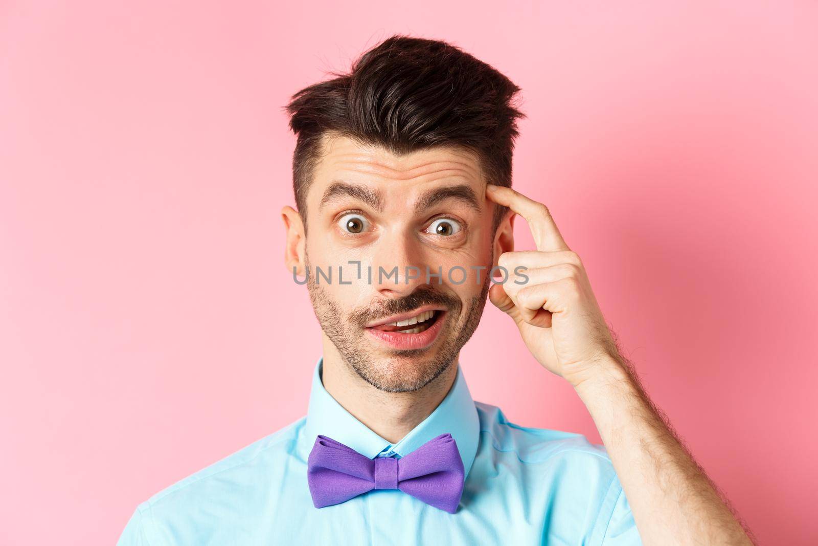 Close-up of confused guy in bow-tie, scratching head and looking puzzled, cant understand something, standing in bow-tie on pink background.