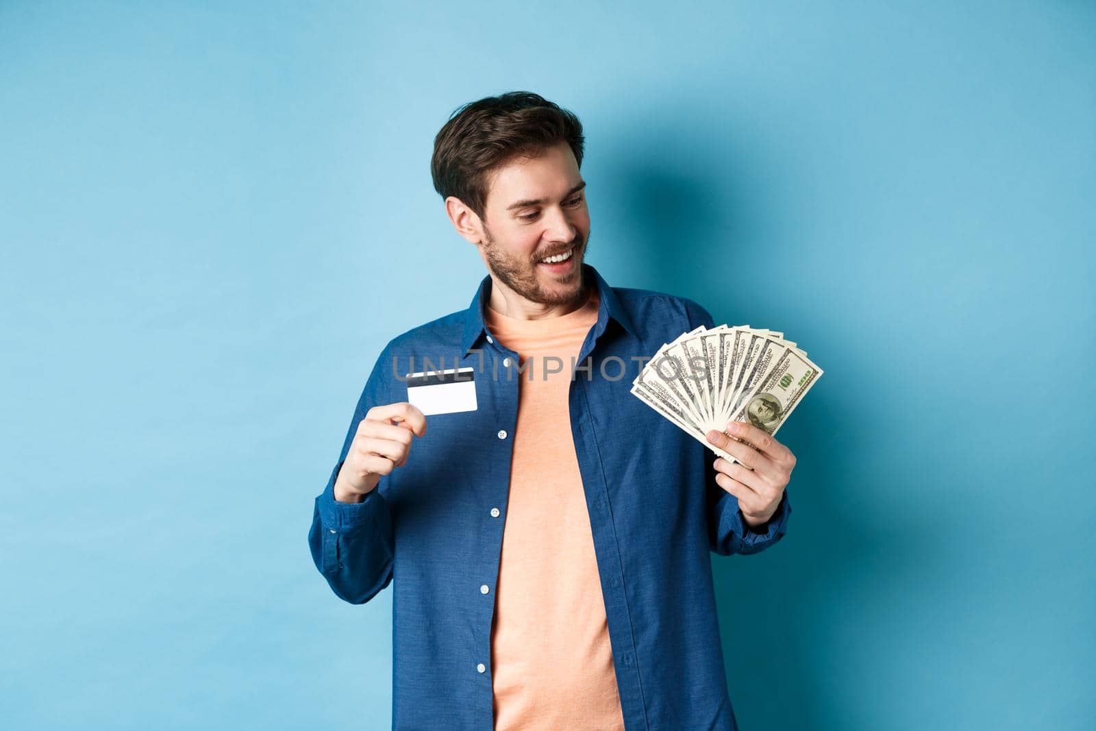 Smiling modern guy looking at cash and showing plastic credit card, standing on blue background. Copy space