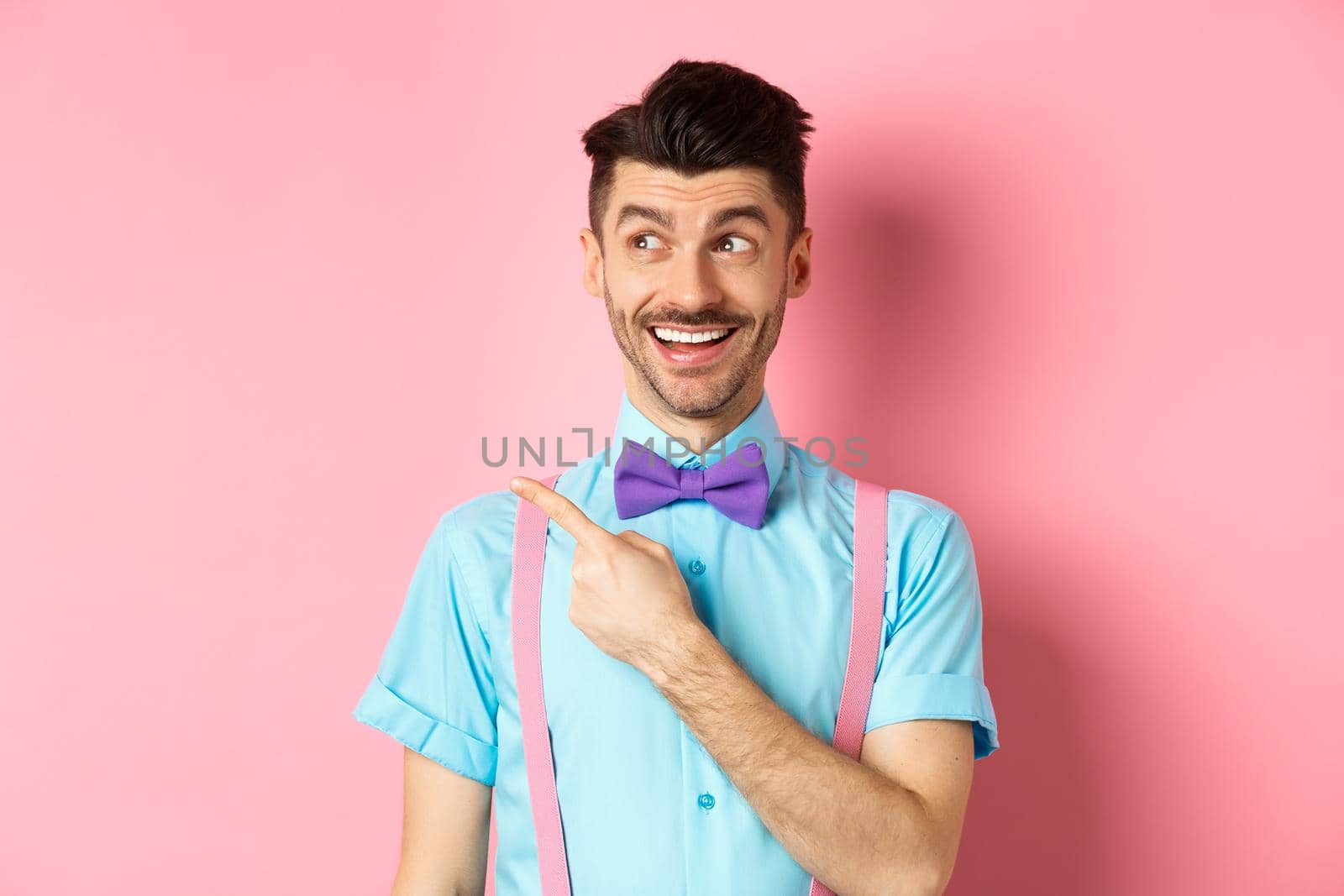 Cheerful caucasian man in funny bowtie looking and pointing hand left, showing promo logo, standing on pink background. Copy space