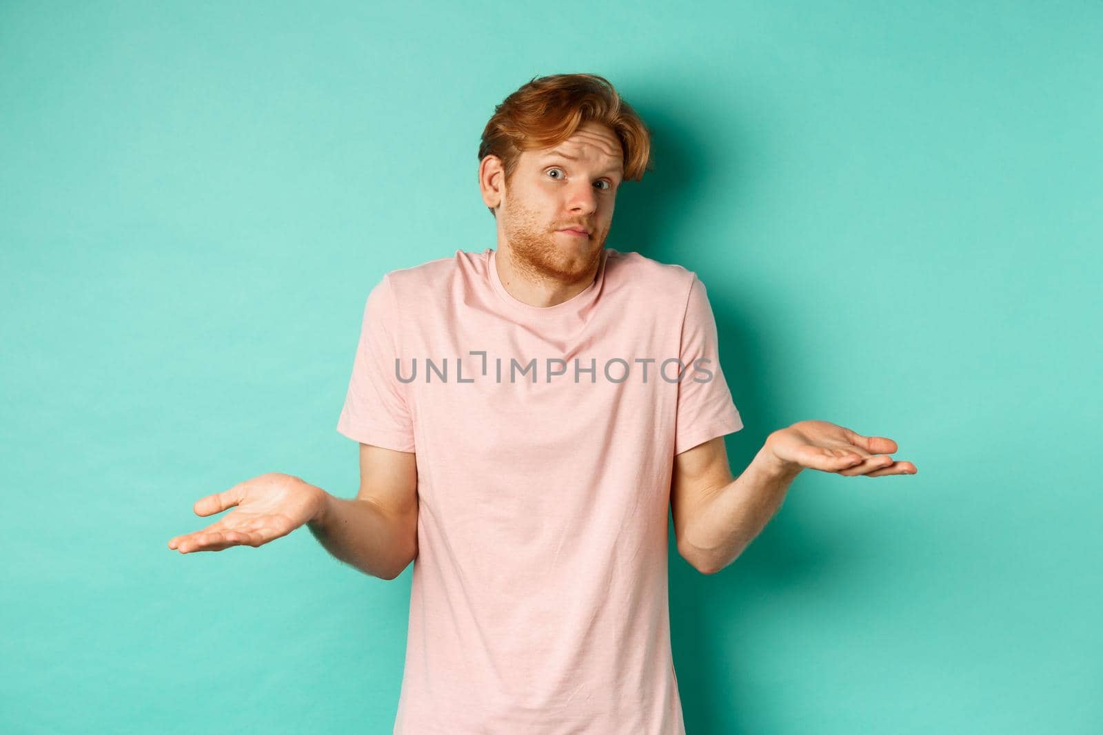 Young redhead guy in t-shirt know nothing, shrugging shoudlers with hands spread sideways, standing confused over turquoise background by Benzoix