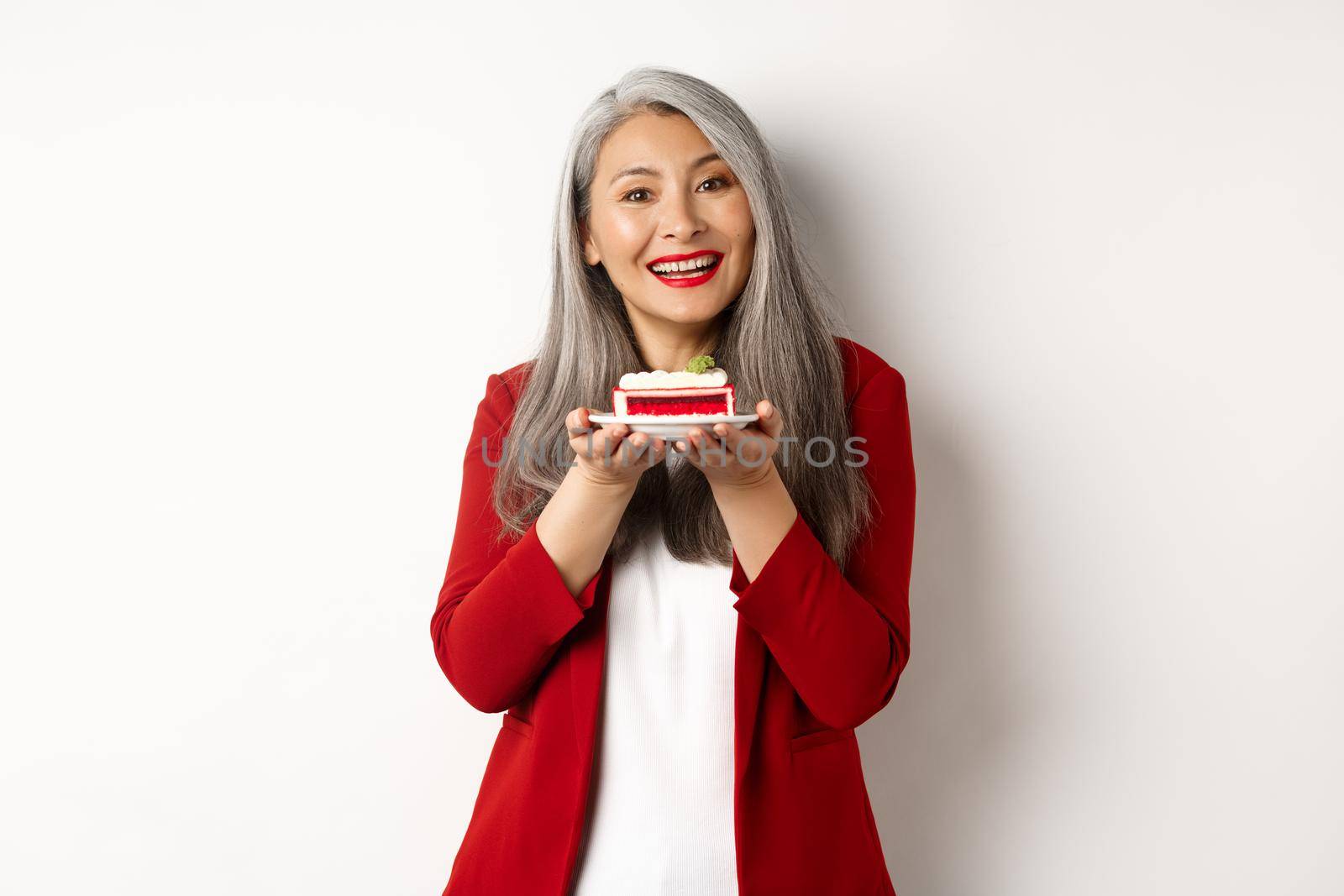 Beautiful asian grandmother holding piece of cake and smiling, standing in red blazer over white background.