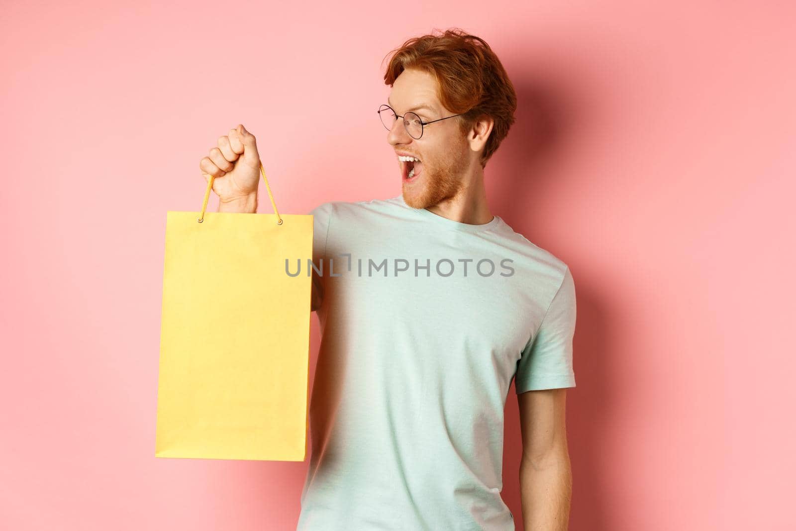 Handsome young man buying presents, holding shopping bag and looking amused, standing over pink background.