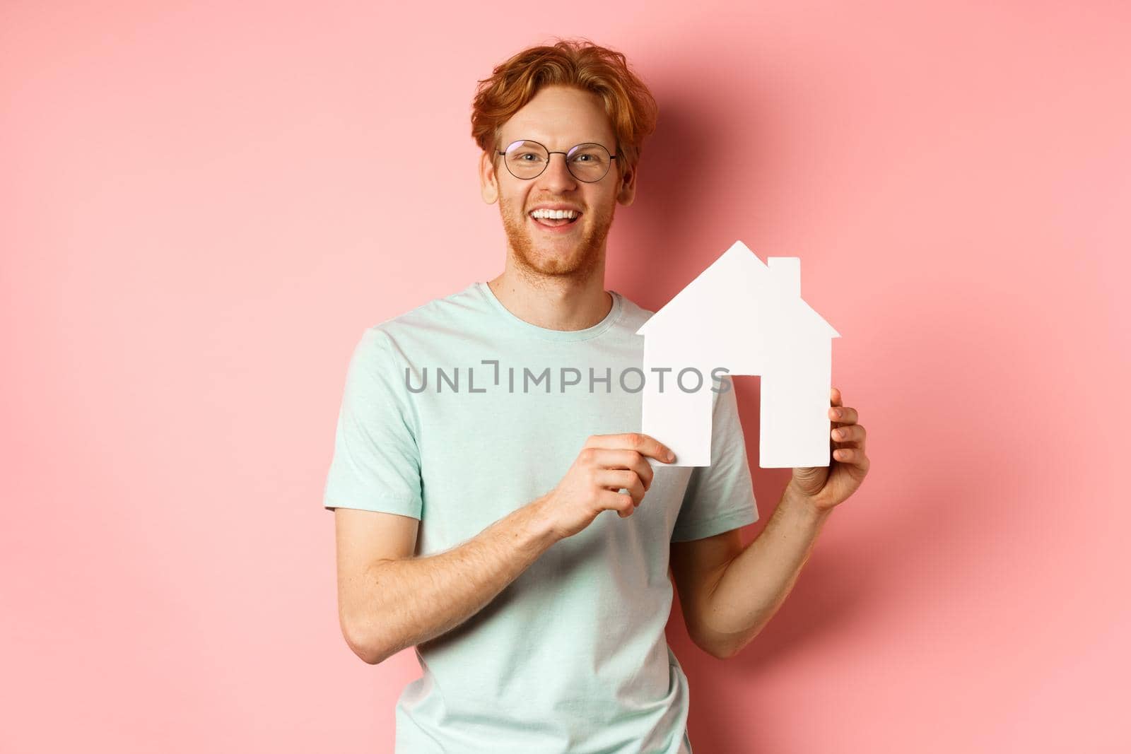 Real estate. Handsome redhead man in t-shirt and glasses, showing paper house cutout and smiling, standing over pink background by Benzoix