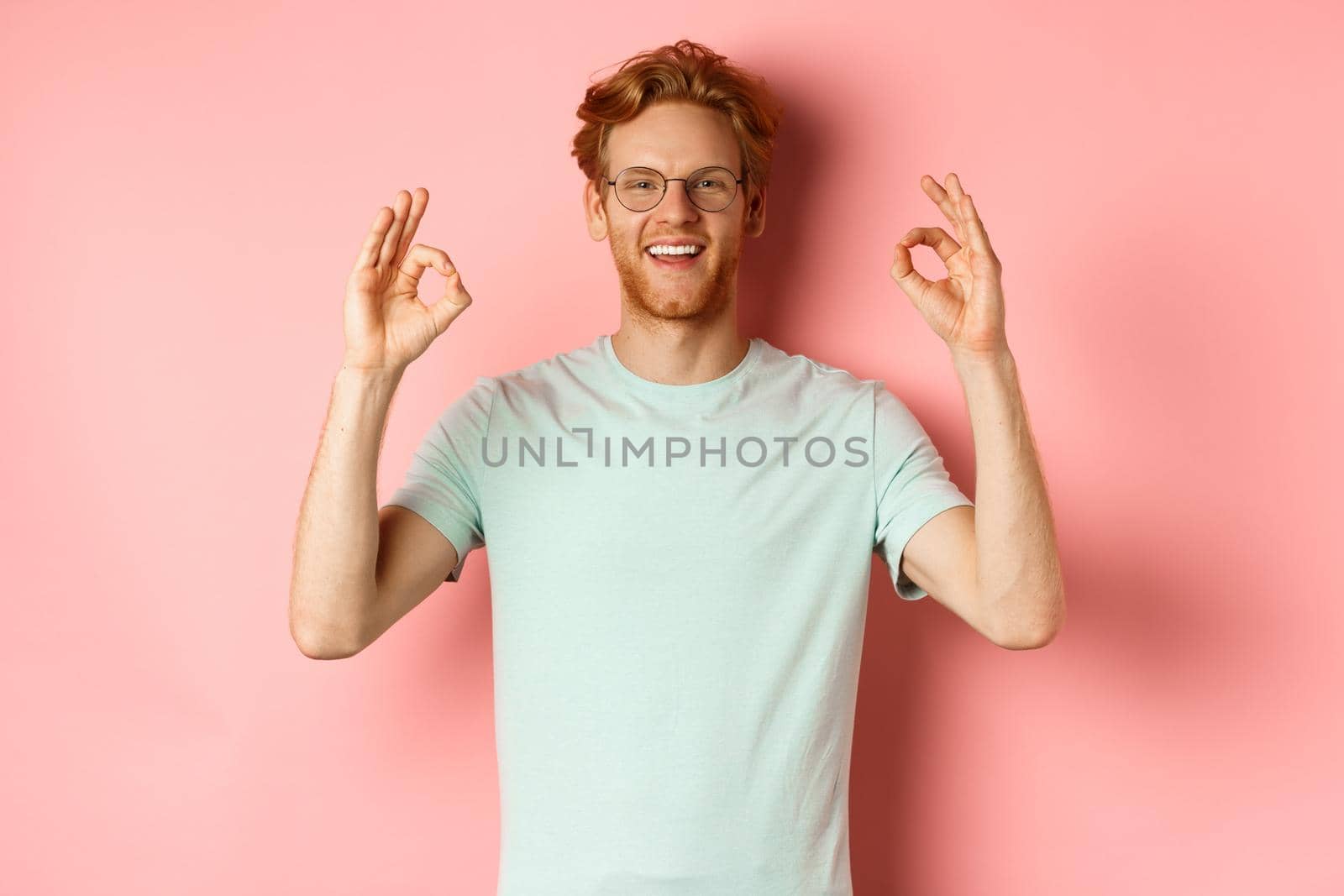Satisfied caucasian man with red hair, wearing glasses and t-shirt, showing okay gestures and smiling, saying yes and approving something, standing over pink background.