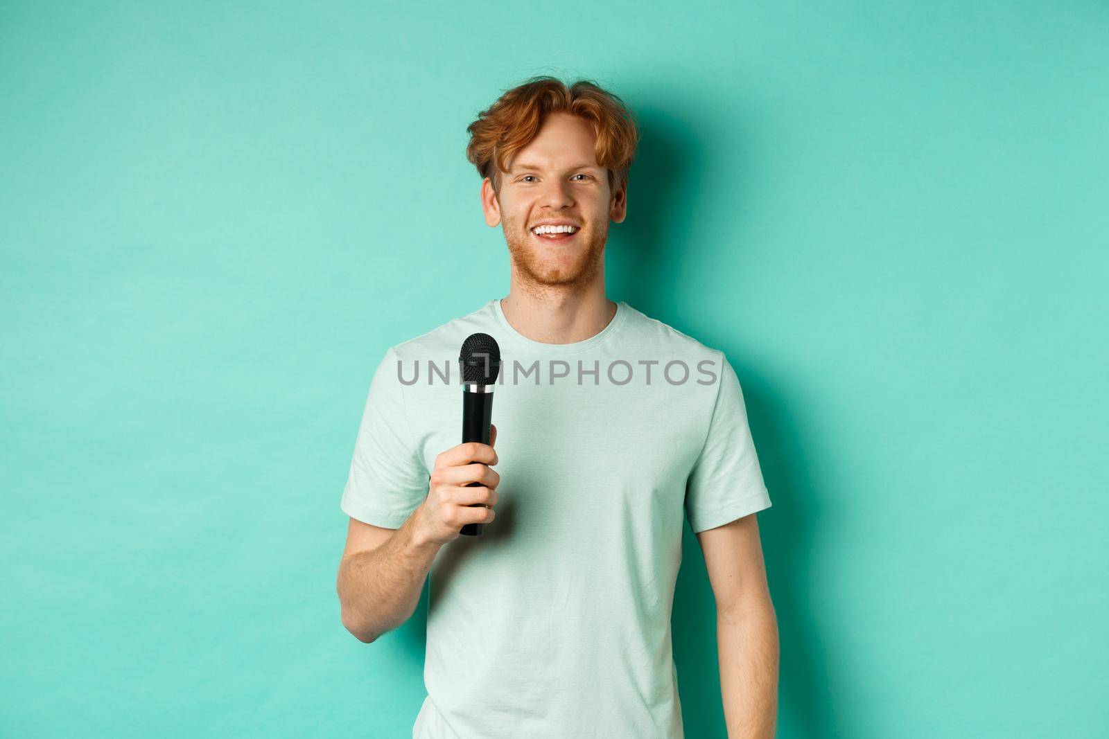 Young redhead man with beard, wearing t-shirt, holding microphone and making speech, singing karaoke, standing over mint background by Benzoix