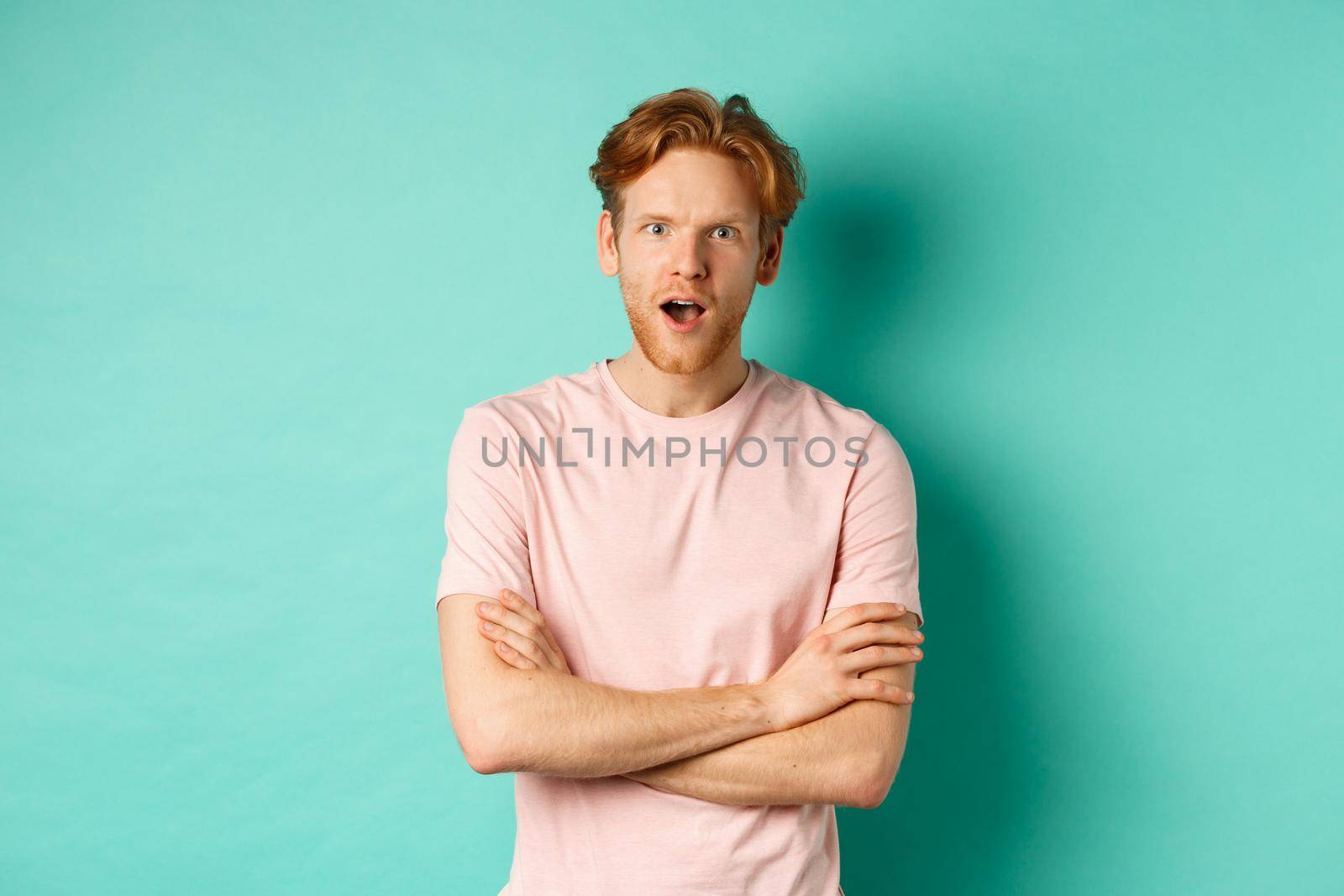 Intrigued redhead guy listening gossips, looking amazed and interested at camera, standing over turquoise background in casual t-shirt by Benzoix