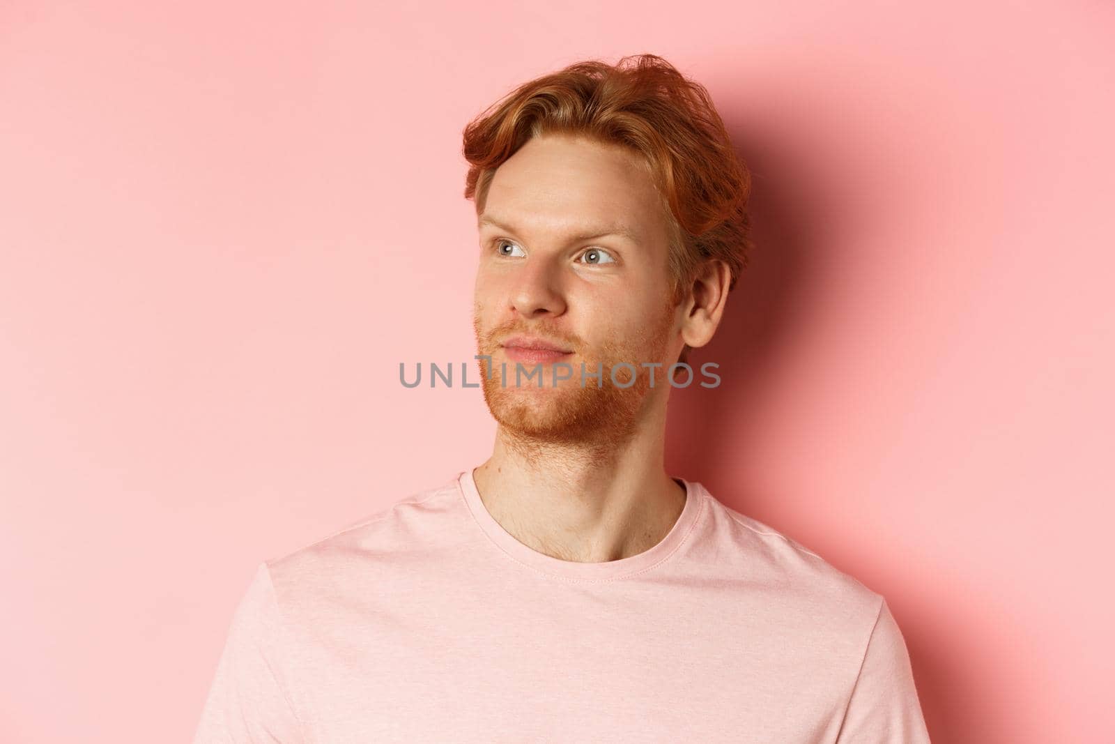 Handsome european male model with red hair and beard, turn head and looking pleased at copy space on left side, standing over pink background by Benzoix