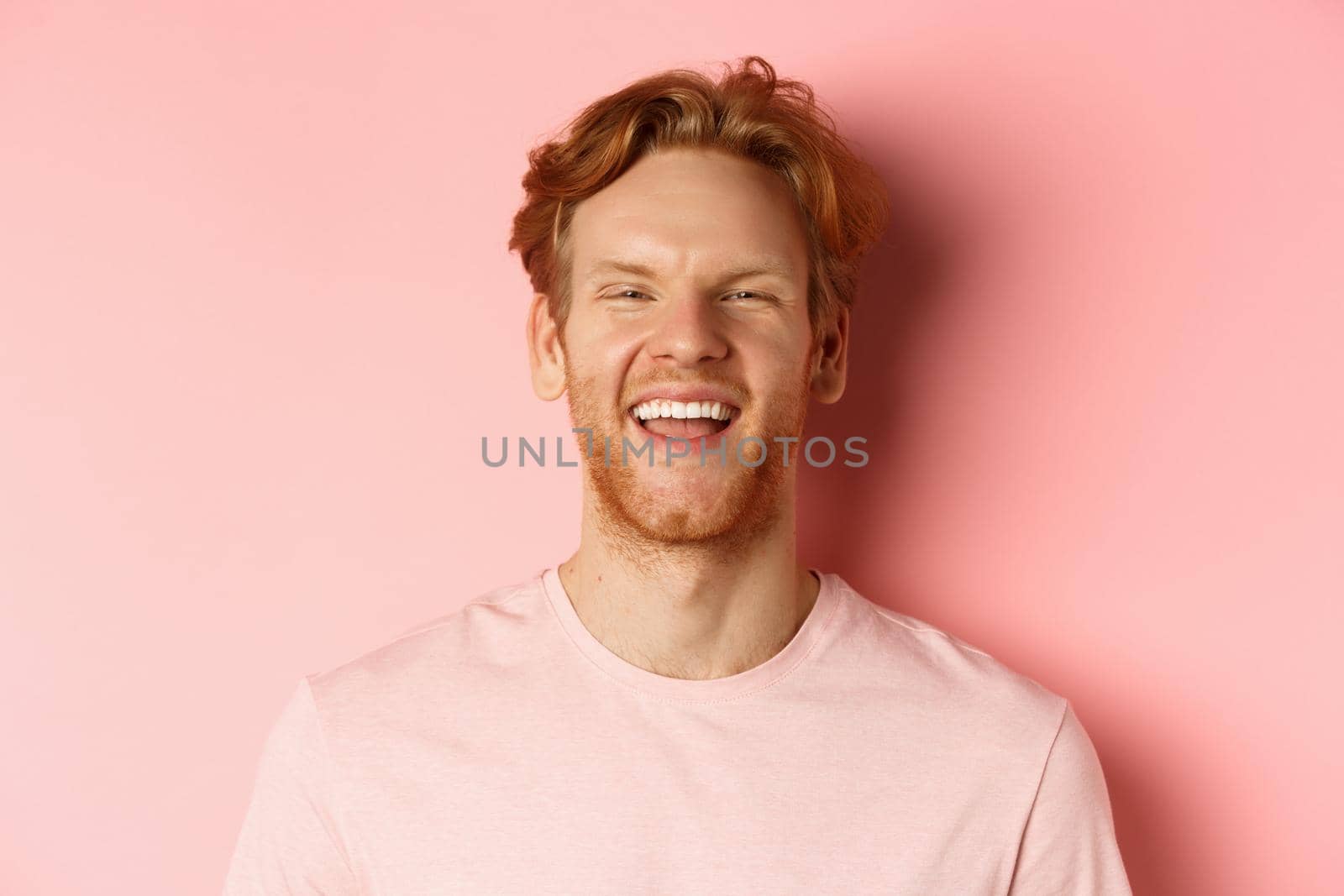 Close up of cheerful young man with messy red hair and beard, laughing and smiling, showing white teeth, looking happy at camera, standing over pink background.