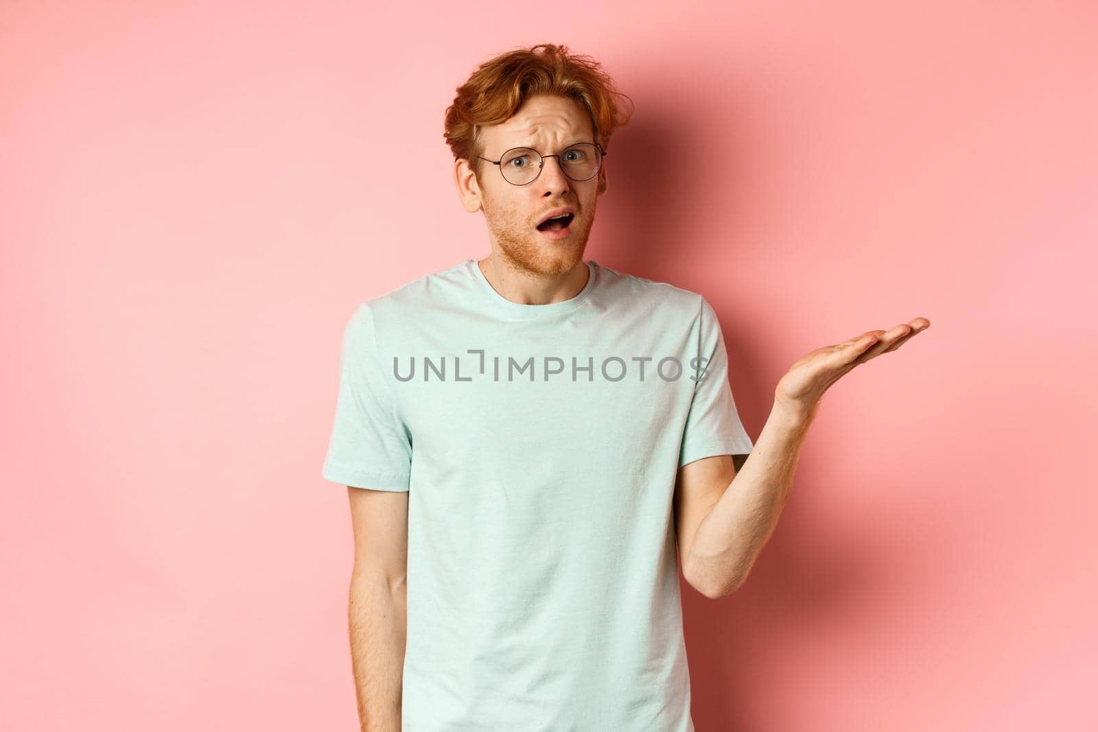 Portrait of handsome redhead man in glasses looking confused, open mouth and stare at camera, cant understand nothing, standing over pink background.