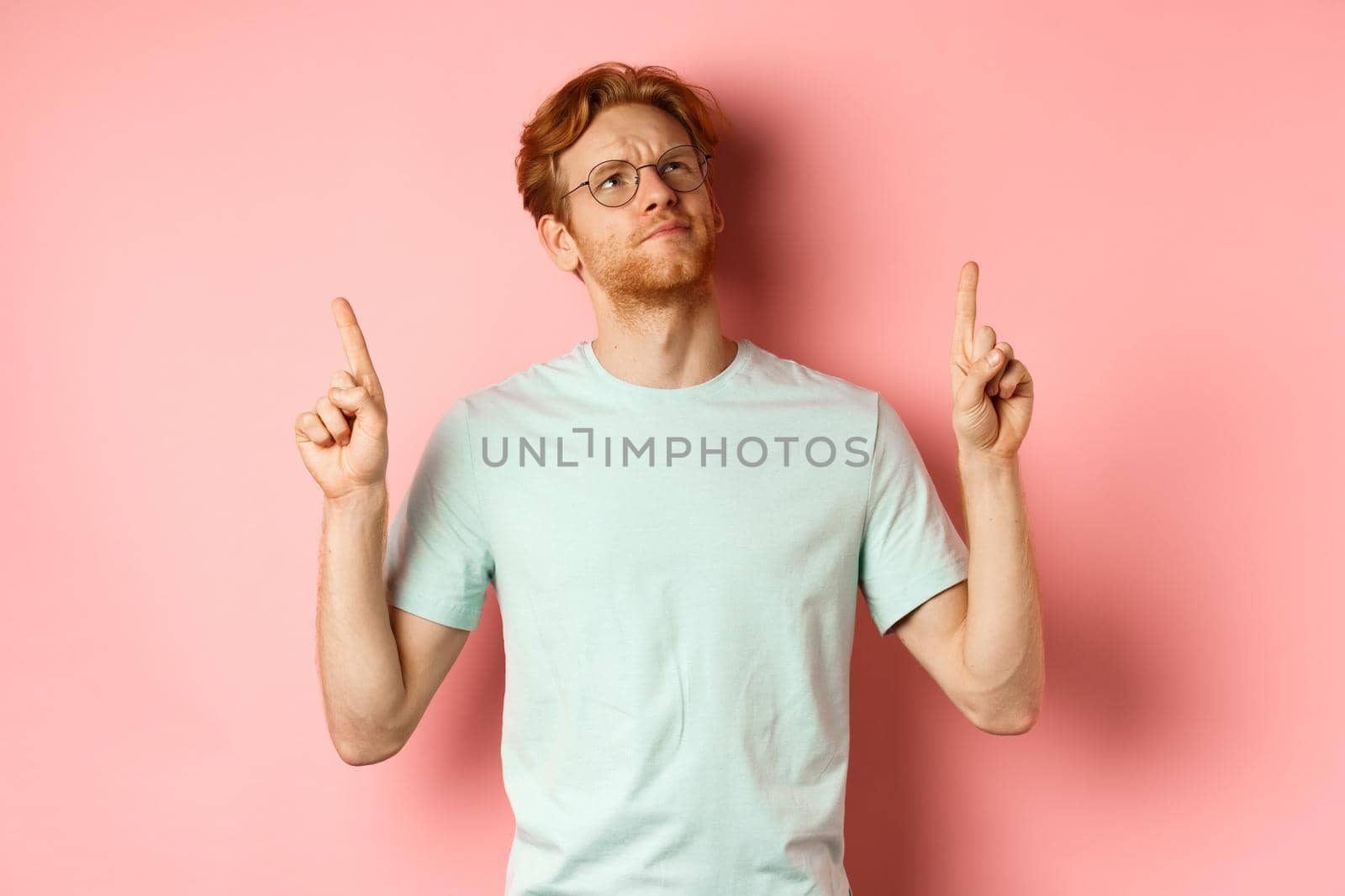 Doubtful redhead guy in glasses frowning, looking and pointing fingers up at something disappointed, standing over pink background by Benzoix