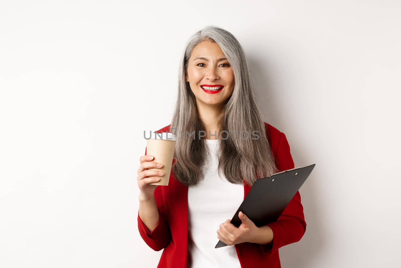 Successful asian businesswoman drinking coffee and smiling, standing with clipboard, wearing red blazer, white background by Benzoix