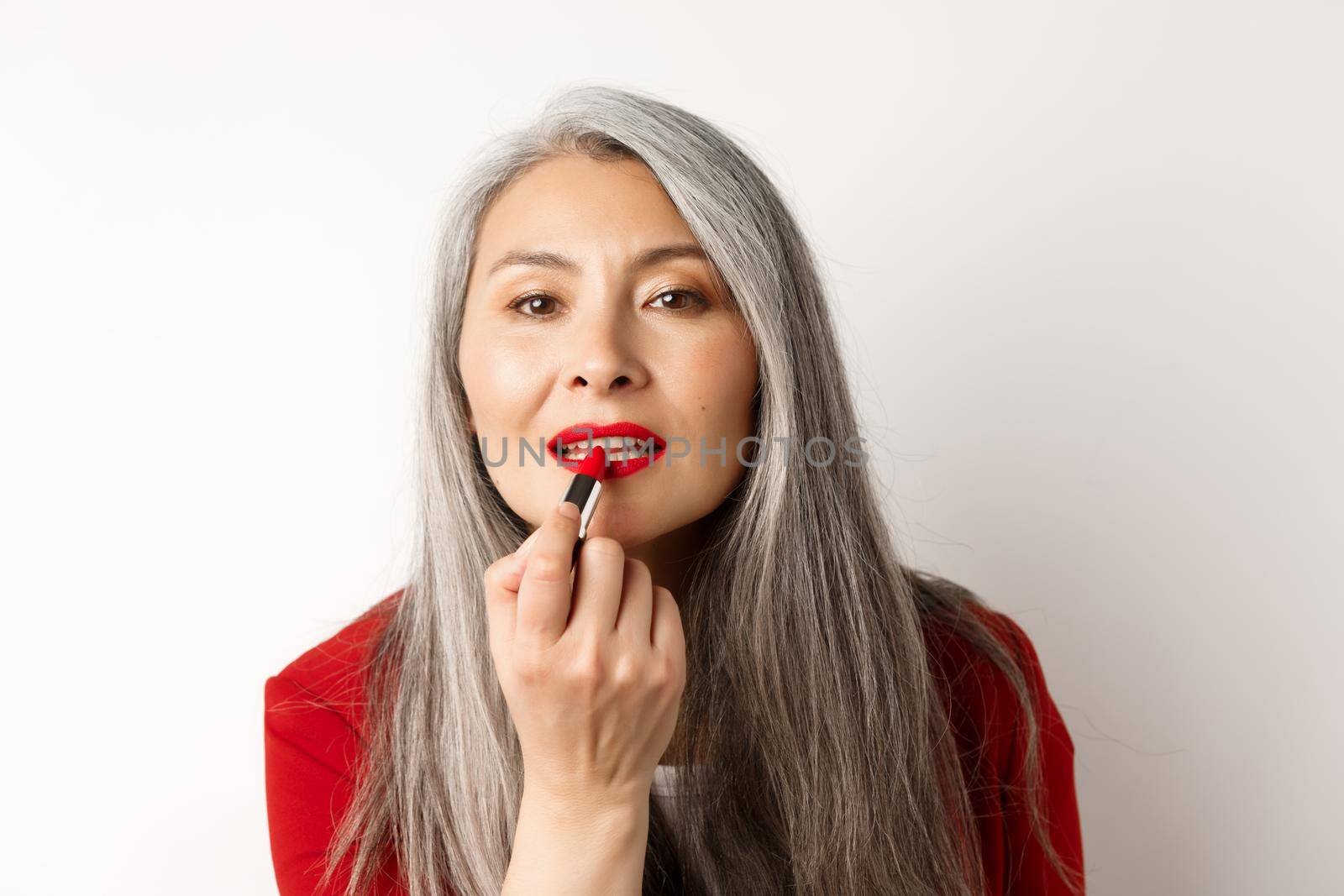 Beauty and fashion concept. Stylish asian mature woman with grey hair, looking in mirror and apply red lipstick, standing over white background by Benzoix