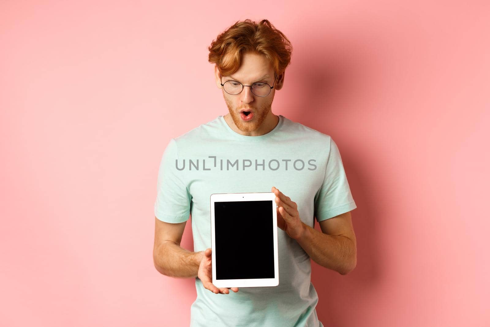 Impressed redhead man in glasses, showing blank digital tablet screen and looking in awe at display, saying wow, standing in t-shirt against pink background.