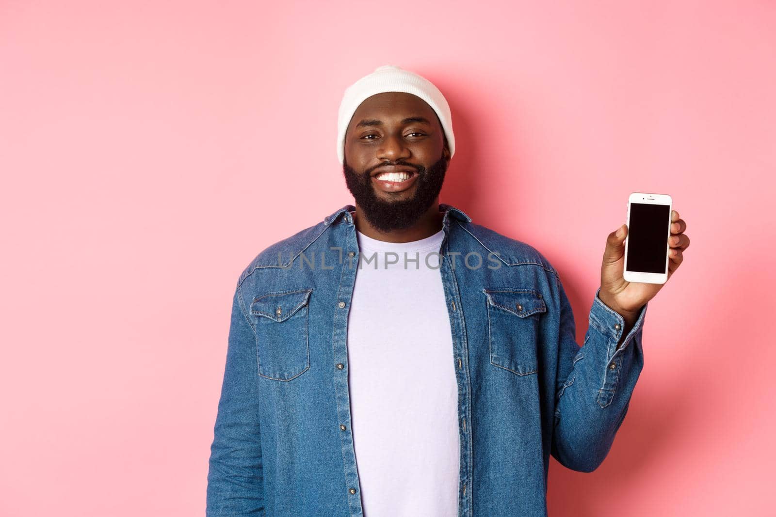 Handsome hipster guy in beanie and denim shirt smiling, showing mobile phone screen with happy face, introduce application, standing over pink background.