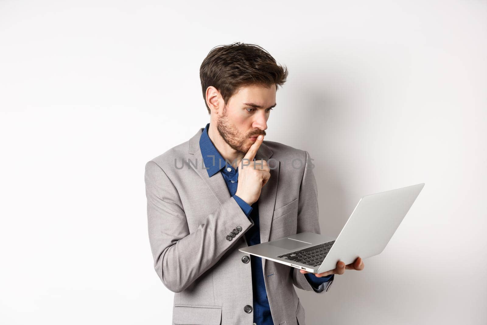 Pensive businessman working on laptop and thinking, looking thoughtful at computer screen, standing in suit on white background.