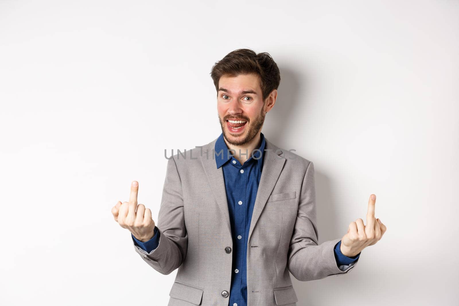 Rude ignorant guy in business suit showing middle fingers and tongue, smiling while mocking people, fuck you gesture, standing on white background.