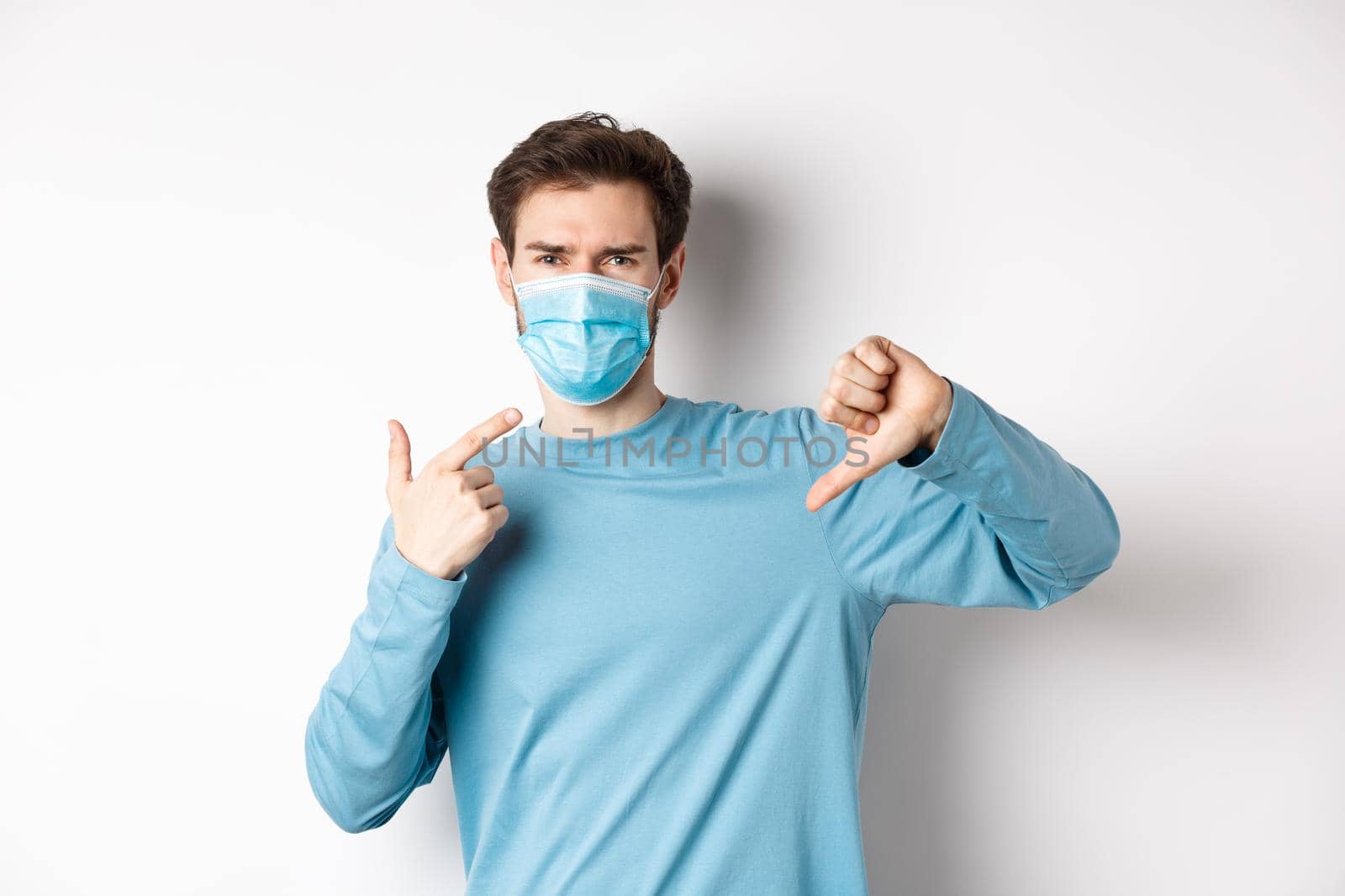 Coronavirus, health and quarantine concept. Angry man pointing at his medical mask and showing thumbs down, complaining on covid, white background.