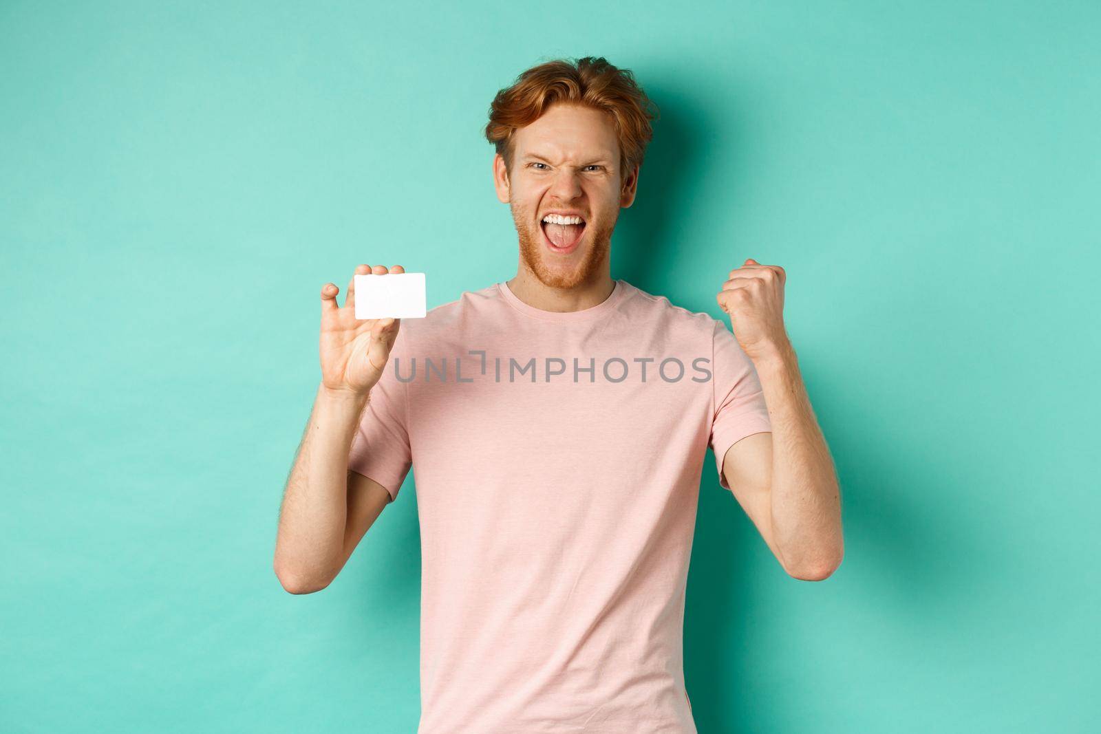 Cheerful young man triumphing, making fist pump to celebrate success, showing plastic credit card, winning prize from bank, turquoise background.