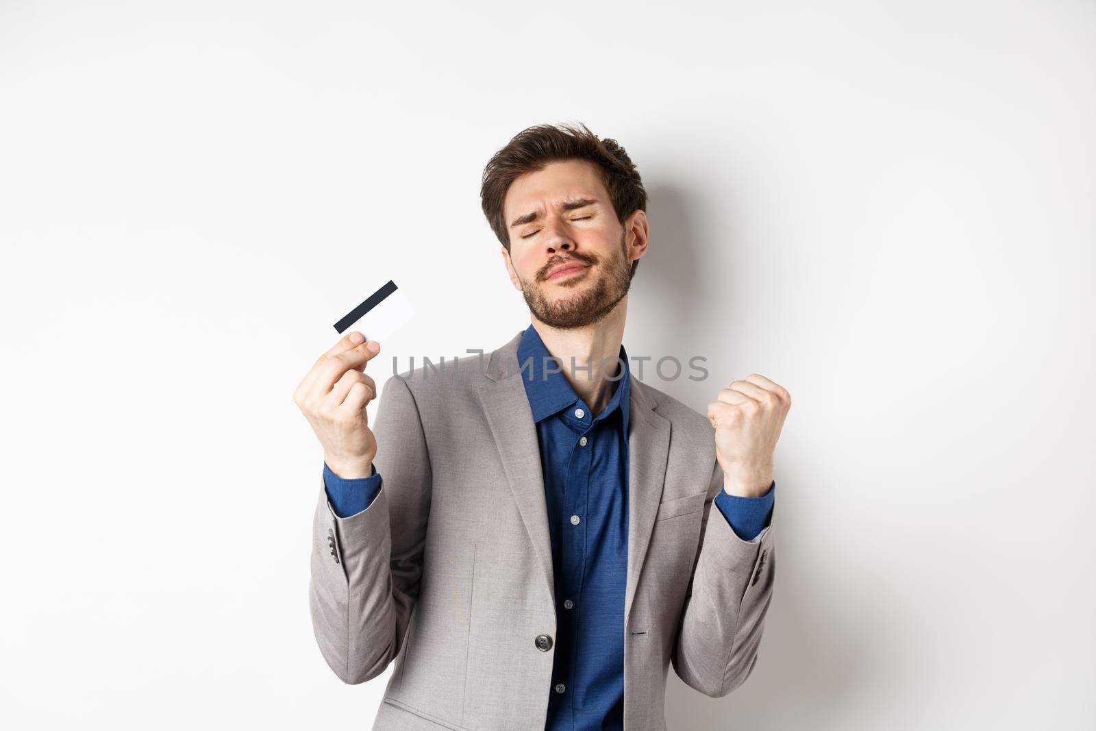 Excited successful businessman dancing with plastic credit card, raising fist pumps and smiling delighted, standing on white background in suit.