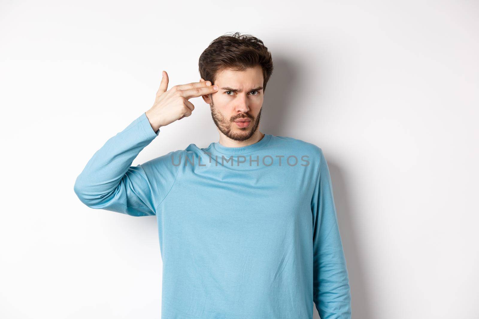 Annoyed young man frowning, showing shooting gun near head gesture, blow brain out sign, standing over white background.